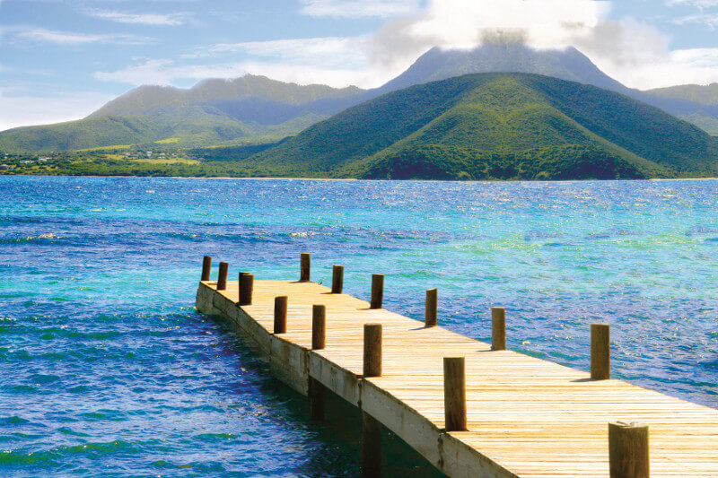 A wooden dock sits with calm blue water and a view of lush mountains.