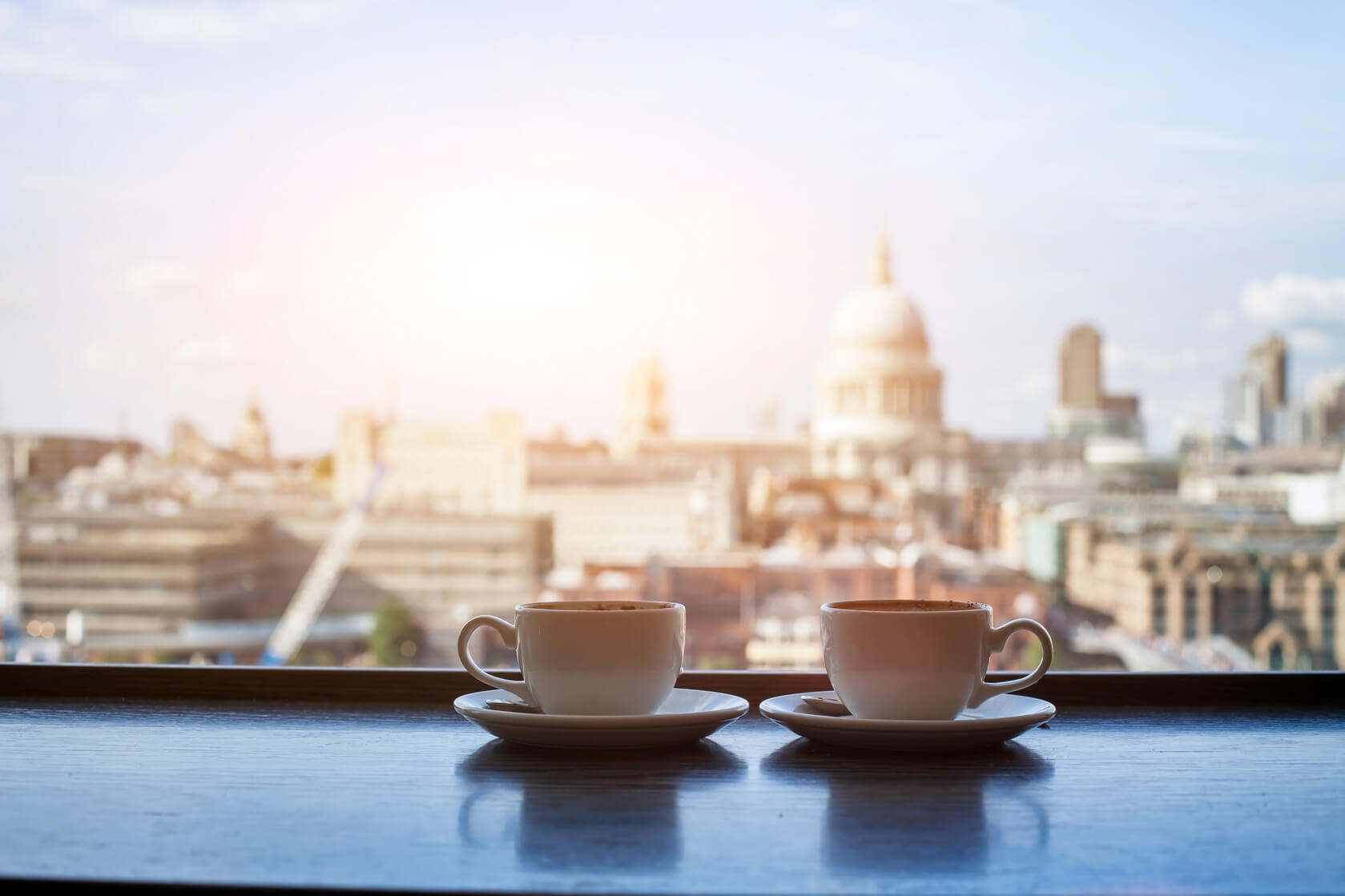Two tea cups sit on a table with the landscape of London behind them. 
