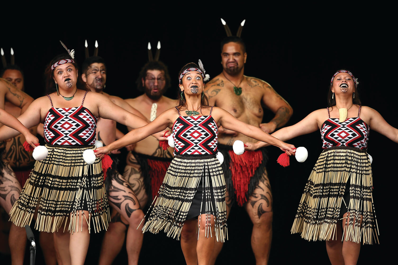 Three women stand in front of a group performing the haka dance